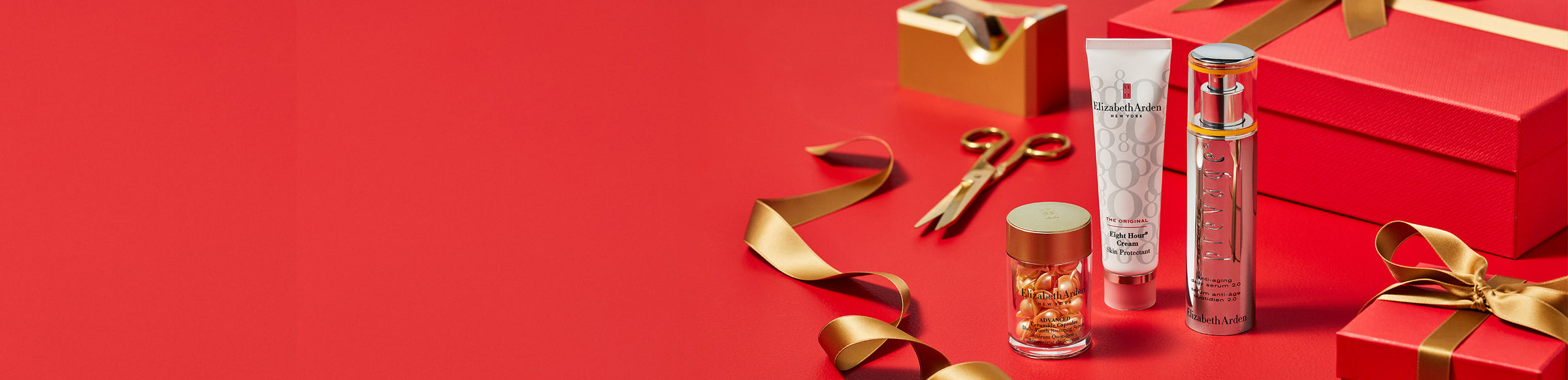 Elizabeth Arden skincare products, Eight Hour Cream Skin Protectant, Daily Youth Restoring Serum Advanced Ceramide Capsules, and Prevage Anti-Ageing Serum 2.0 arranged on a red background with gold ribbon, scissors, and festive gift boxes.