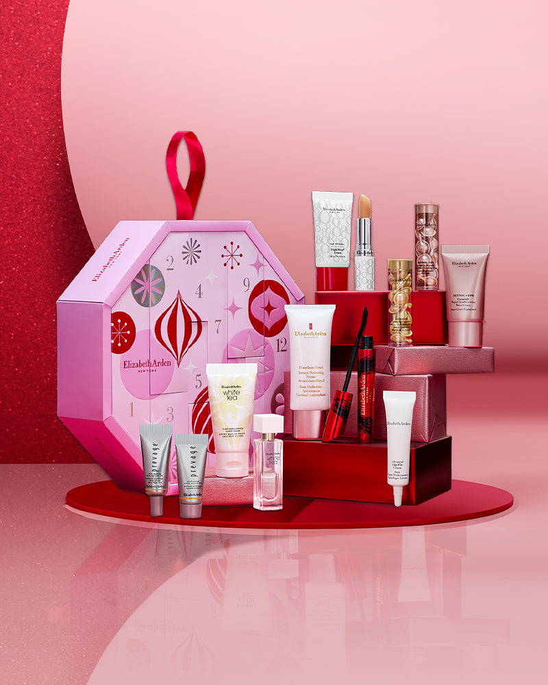 A cosmetics gift set displayed on a red circular pedestal against a pink and red studio background. The set includes a pink patterned gift box with a red loop handle, several red and white tubes, bottles, and jars, and a small red box.