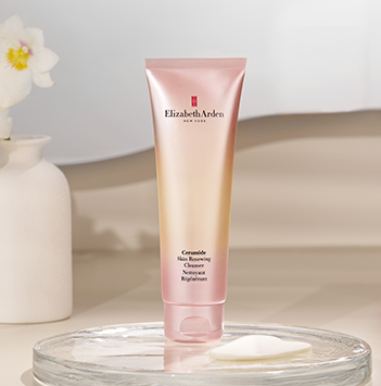 Elizabeth Arden Granulate Skin Boosting Serum in a metallic rose gold squeeze tube, displayed on a clear glass tray with a white cotton pad next to a white ceramic vase with a flower.