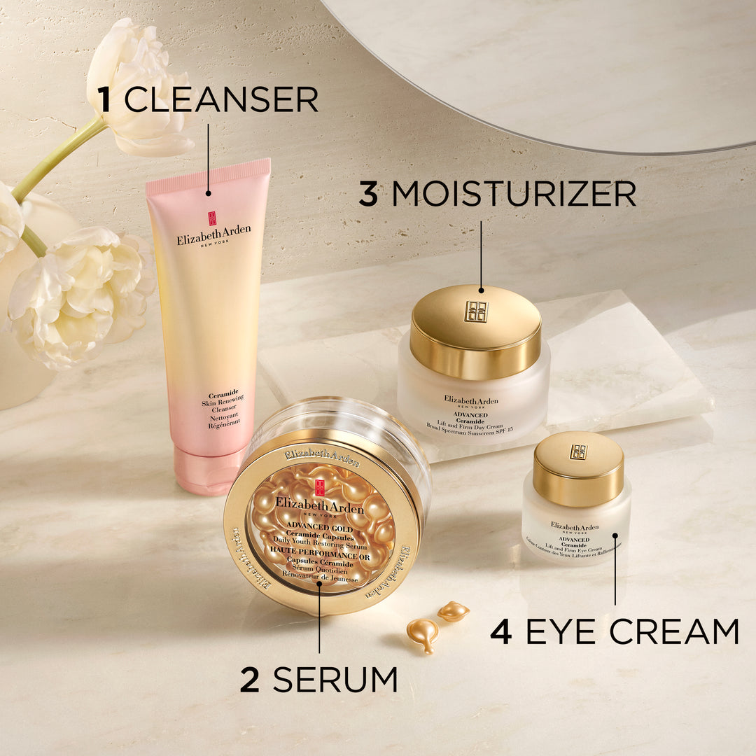 Four Elizabeth Arman skincare products arranged on a light marble surface with labeled callouts: cleanser, serum, moisturizer, and eye cream. The products include a pale pink cleanser tube, a clear jar with gold lid showing gold-toned serum inside, a white moisturizer jar with gold lid, and a small white eye cream jar with gold lid.