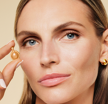 Close-up of a woman holding a small gold beauty dropper near her cheek, with a soft beige background.