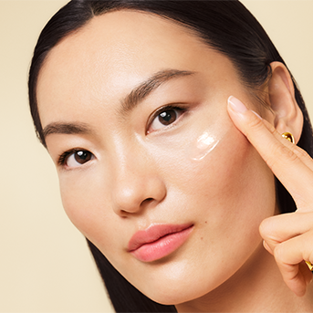 Close-up of a woman applying a glossy, pearlescent skincare cream or serum to her cheek with her index finger. She has dark hair pulled back, and a small gold earring is visible.