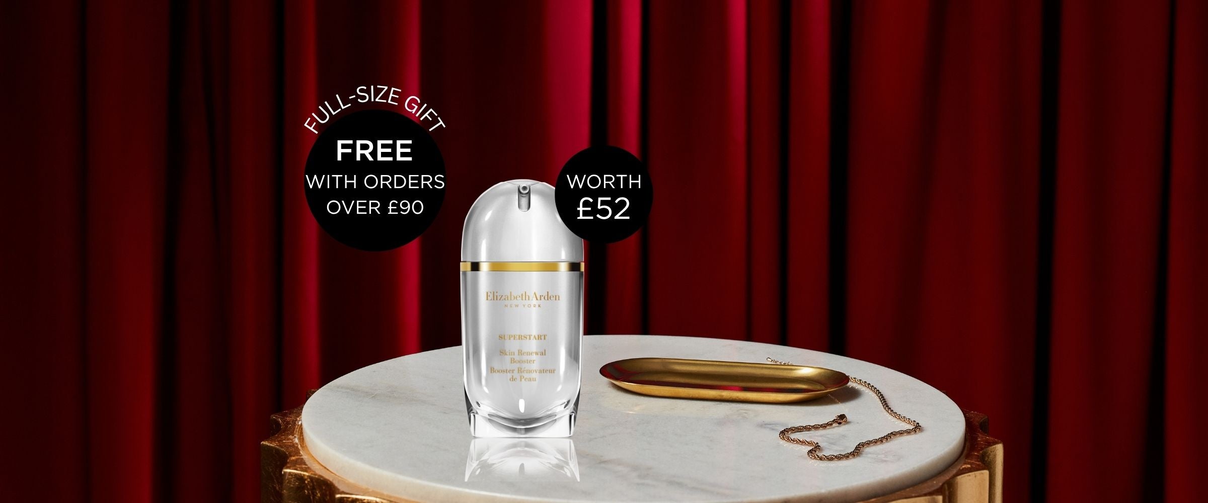 Elizabeth Arden Superstart Skin Renewal Booster placed on top of a circular marble top with a small gold long plate next to it and emphasized against a deep red curtain background. 