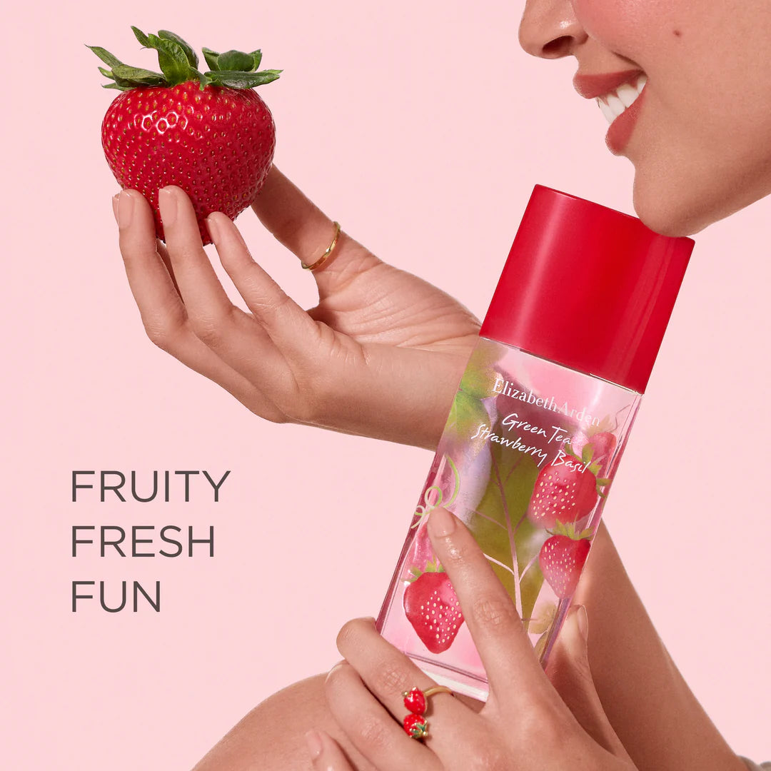 Person holding a strawberry and a bottle of Elizabeth Arden Green Tea Strawberry Beret perfume against a pink background.