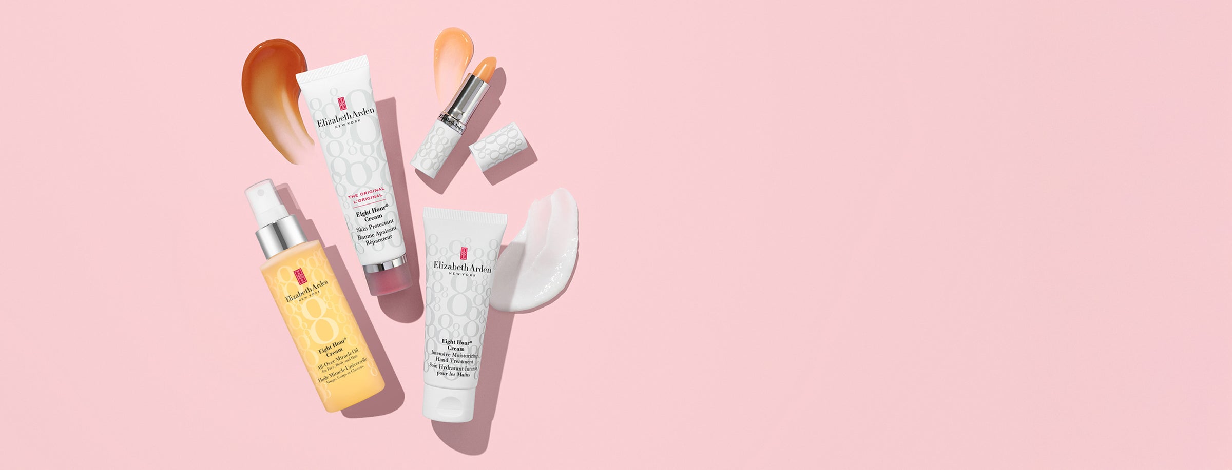 Four Elizabeth Armand skincare products arranged on a solid pink background: a yellow serum bottle, a pink cream tube, a white cream tube, and a lip product with its cap removed. White cosmetic sponges and orange makeup smears are placed around them.