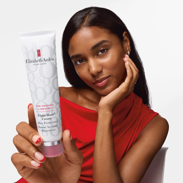 A woman in a red top holds up a tube of Elizabeth Arden Eight Hair Cream against a clean white background.